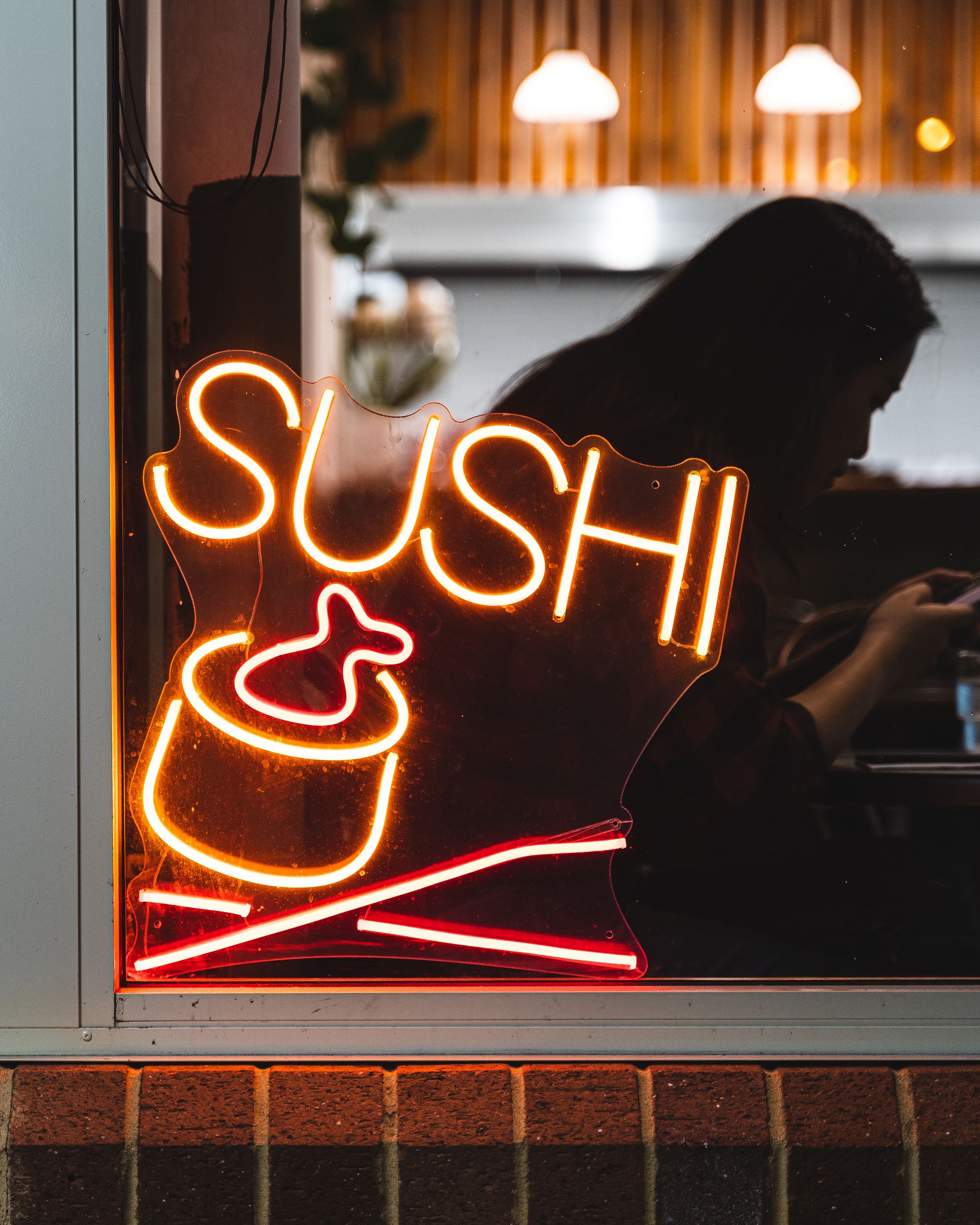A neon lit sign that says "sushi" showing a maki roll with a fish sticking out and chopsticks underneath it