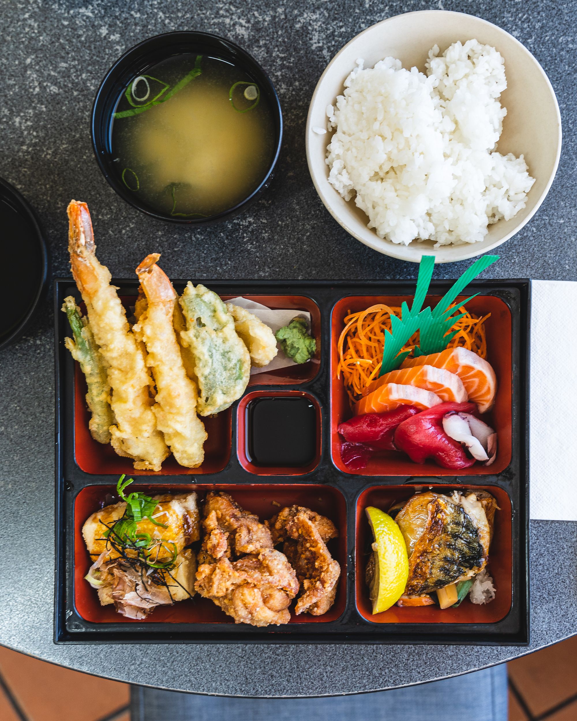Top down of a bento box with lots of Japanese dishes, a bowl of rice in the back and a bowl of miso soup