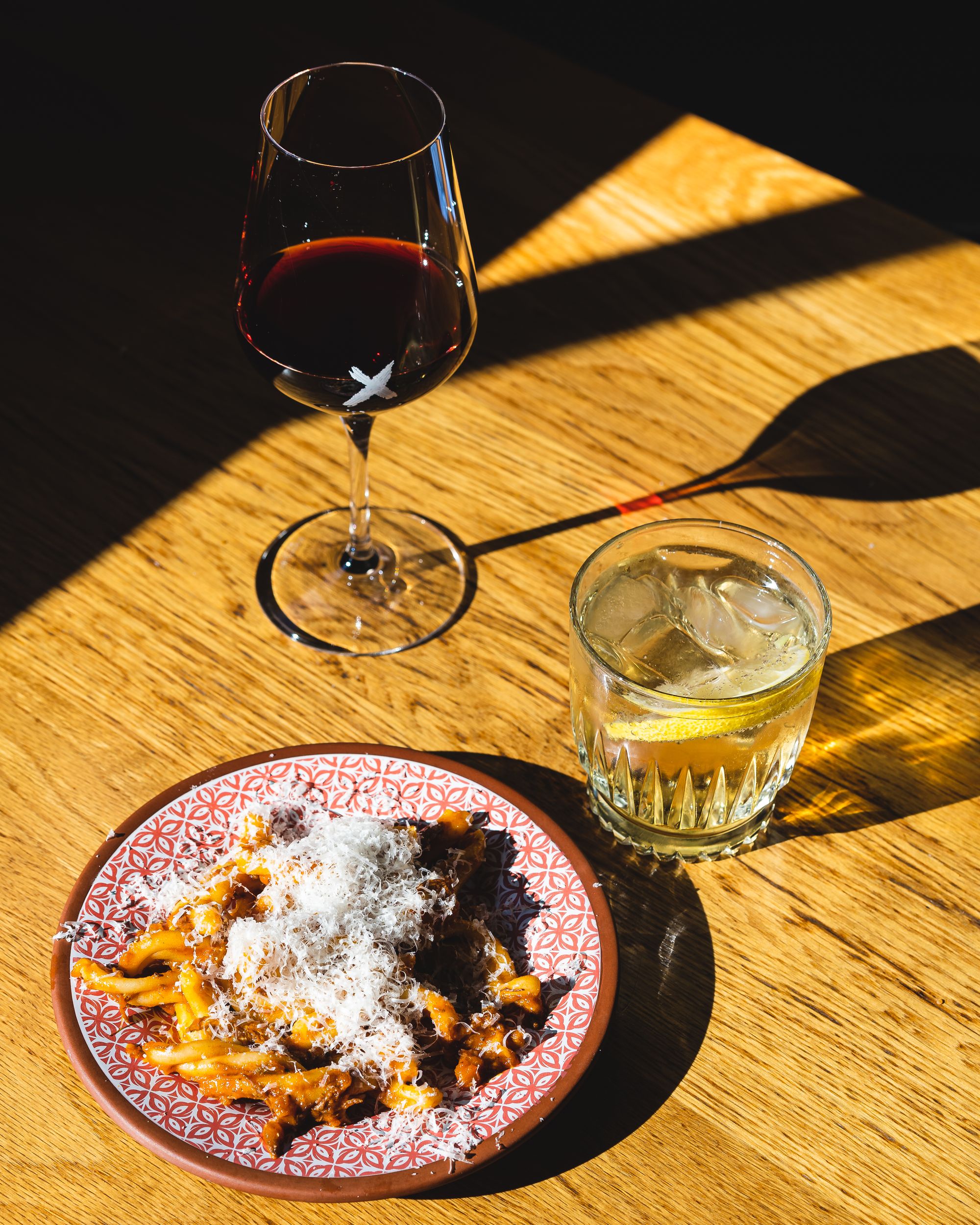 Harshly lit shot of pasta, a mocktail class and wine glass with long casted shadows from the harsh light