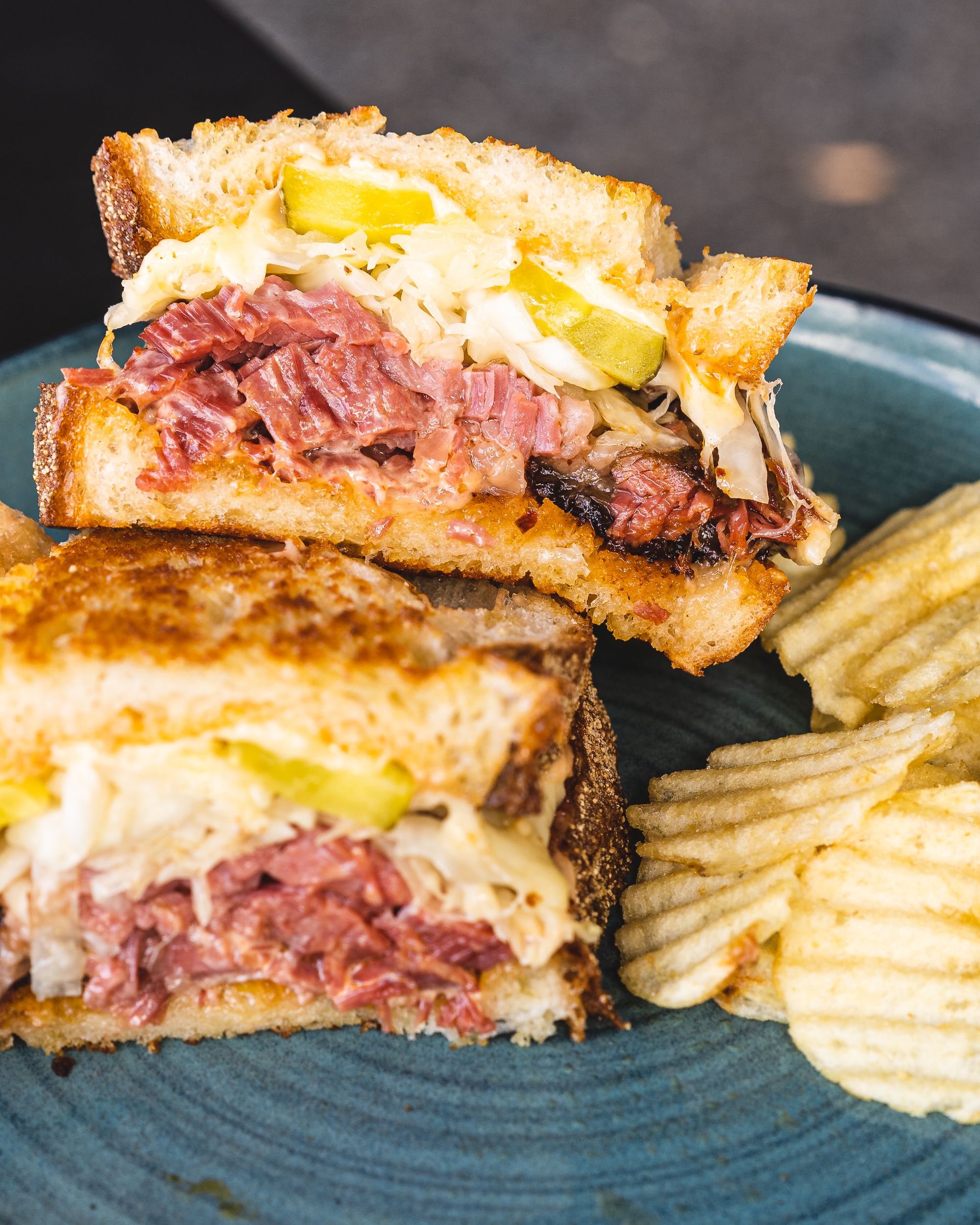 Close up of Reuben sandwich with pastrami, cheese, pickle and a side of crisps