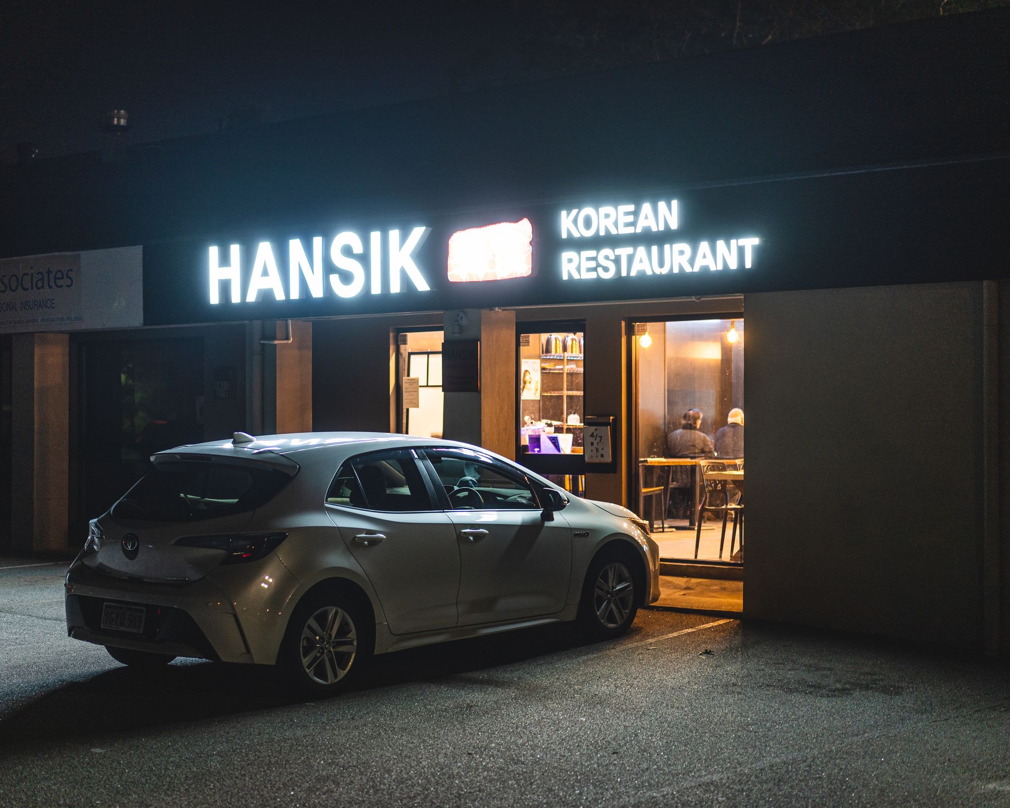 Exterior shot of a restaurant with a car outside, illuminated by the restaurant sign and the restaurant interior