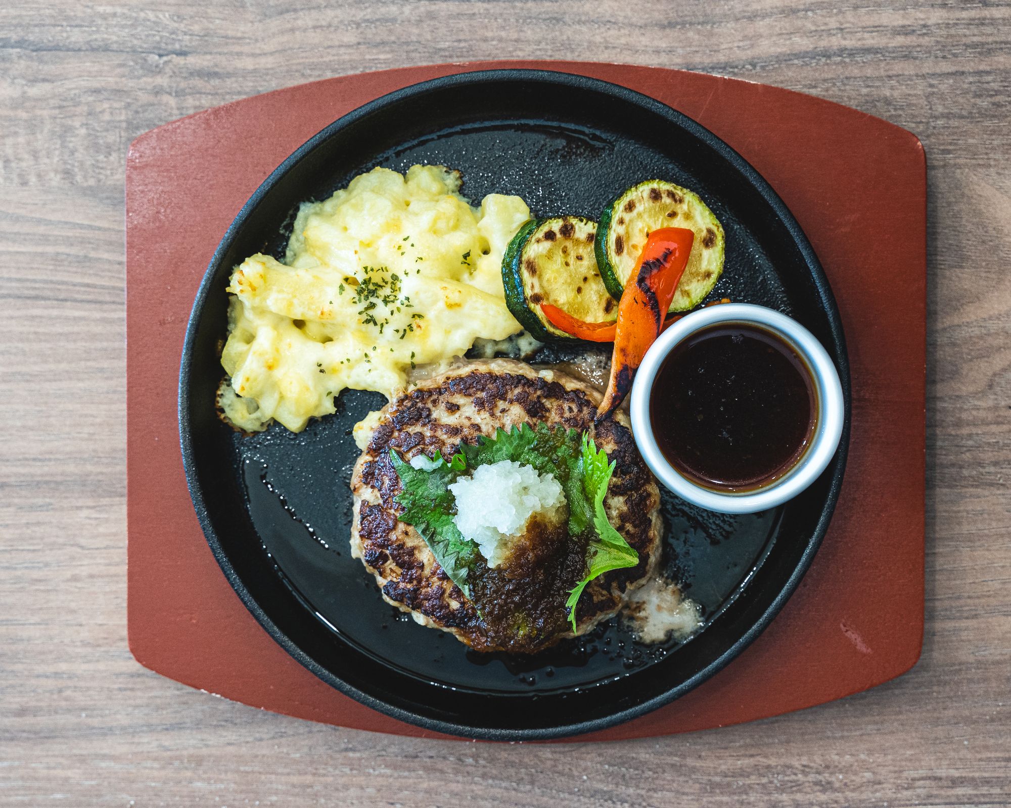 Japanese hamburg steak with potato gratin and roasted vegetables, sitting ontop of a sizzling metal pan
